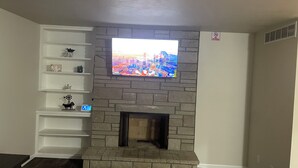 A living room with a stone fireplace, a mounted TV, and a white shelving unit with decorative items. - Charming 2-bedroom house in Jefferson City with AC (Jefferson City)