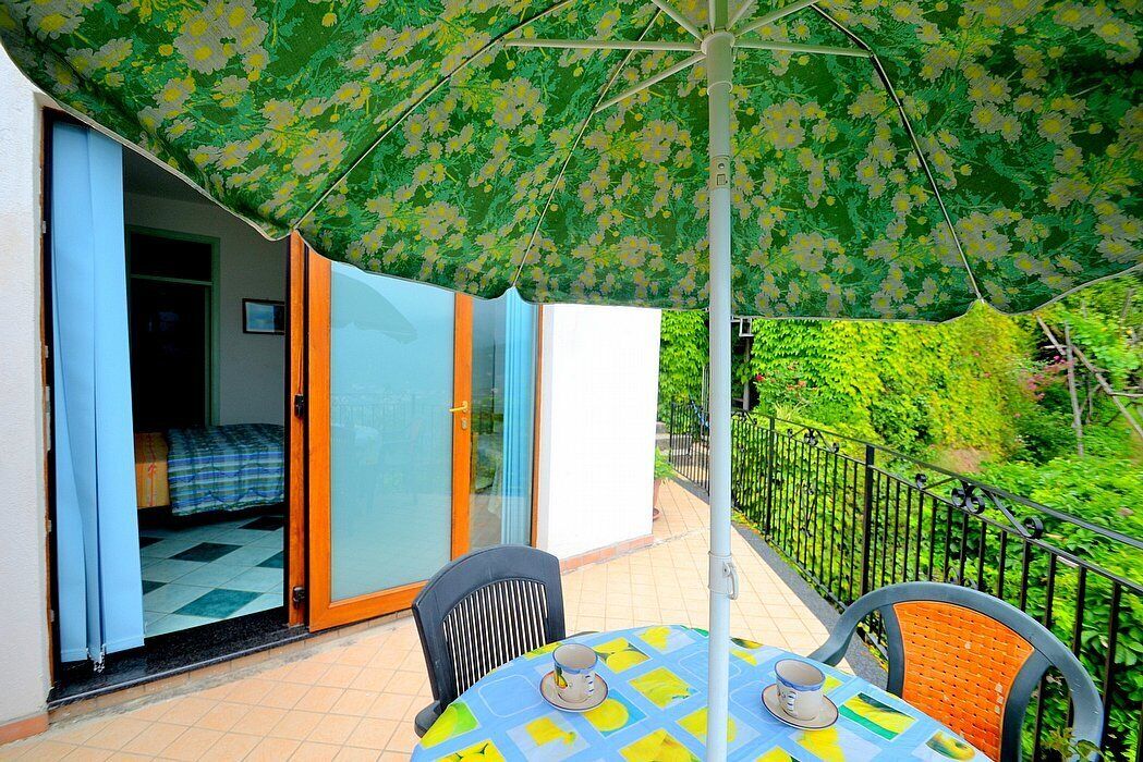 A balcony with a table and chairs, a green floral umbrella, and a view of greenery.