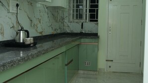 A kitchen with green cabinets, a marble countertop, a kettle, and a window with blinds. - Serene Legacy Park View (Mysore)