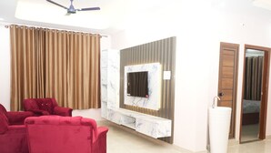 A modern living room with a flat-screen TV mounted on a marble wall, red sofas, a ceiling fan, and a bathroom with a sink and mirror. - Serene Legacy Park View (Mysore)