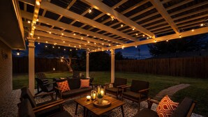 Un patio al aire libre con una pérgola, luces colgantes y mobiliario exterior.