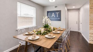 Un comedor con una mesa de madera dispuesta para cuatro personas, sillas traslúcidas, un centro de mesa decorativo y un letrero en la pared que dice 