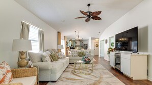 A spacious living room with a sofa, coffee table, and a ceiling fan. A kitchen area with a refrigerator and oven is visible in the background. - Private Pool & Golf Cart! Villa By Spanish Springs (The Villages)