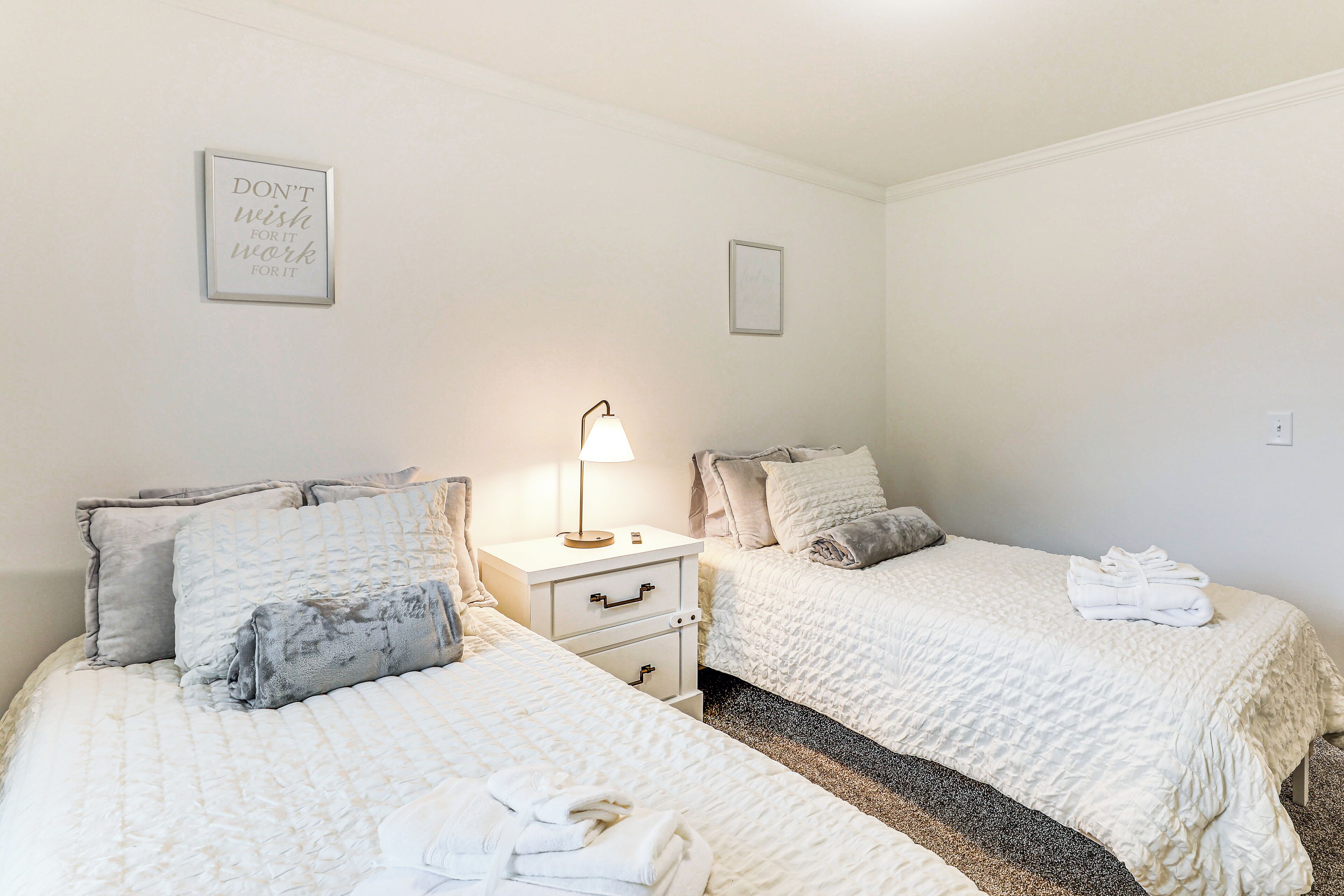 A hotel room with two beds, a nightstand, a lamp, and a motivational quote on the wall.