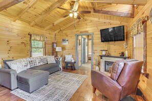Interior acogedor de una cabaña con sofá, reposapiés, sillón de cuero, televisor, chimenea y ventilador de techo.