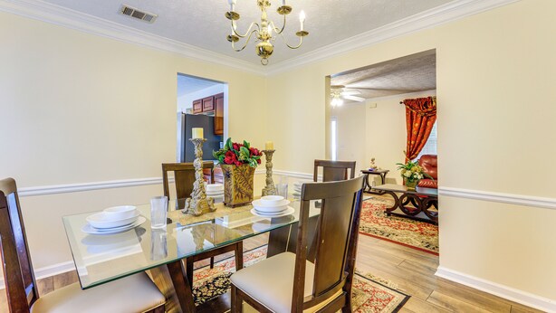 Uma sala de jantar com mesa de vidro, cadeiras de madeira, um candelabro e vista para uma sala de estar com cortina vermelha.