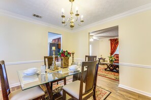 Un coin repas avec une table en verre, des chaises en bois, un lustre et une vue sur un salon agrémenté d’un rideau rouge.