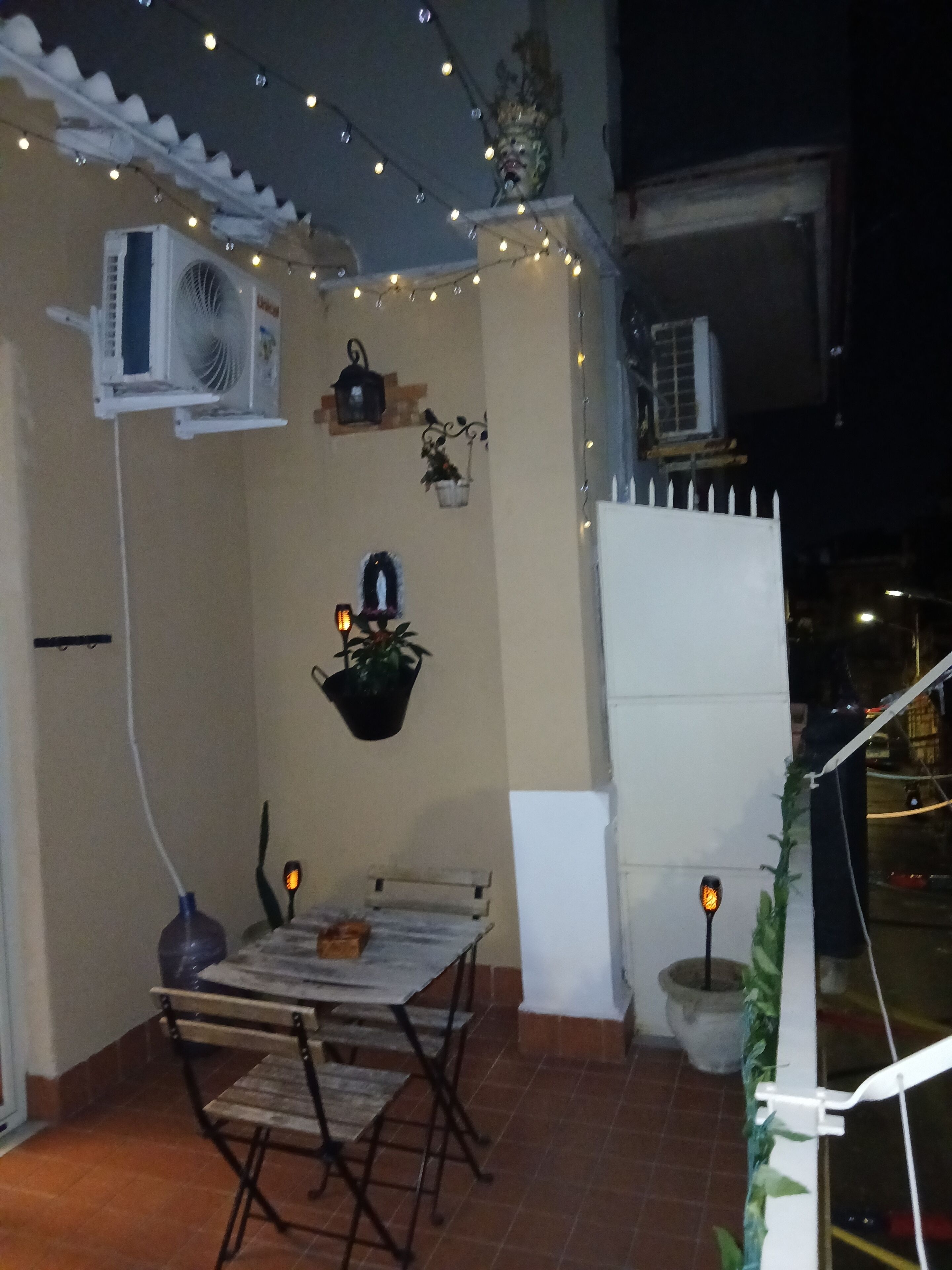An outdoor patio with a table and chairs, string lights, potted plants, and an air conditioning unit.