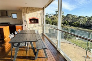 Un balcón con una mesa y sillas de madera, una chimenea de piedra y vistas a un río y árboles.
