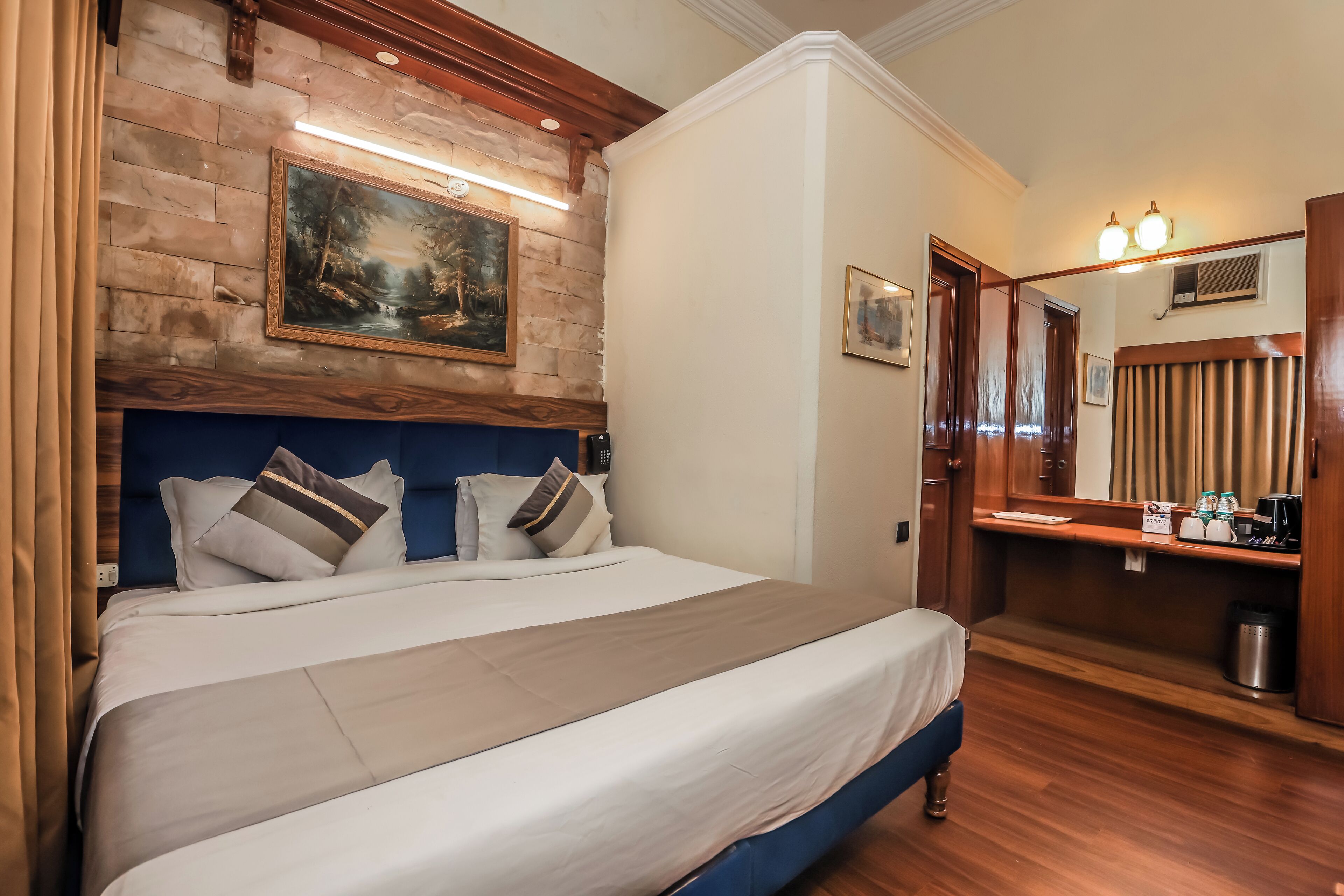 A hotel room with a large bed, a wooden headboard, a painting above the bed, a desk with a mirror, and a small trash bin.