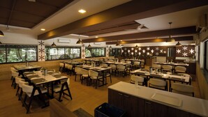 A spacious restaurant with multiple dining tables and chairs, large windows with a view of greenery, and a modern ceiling design.
