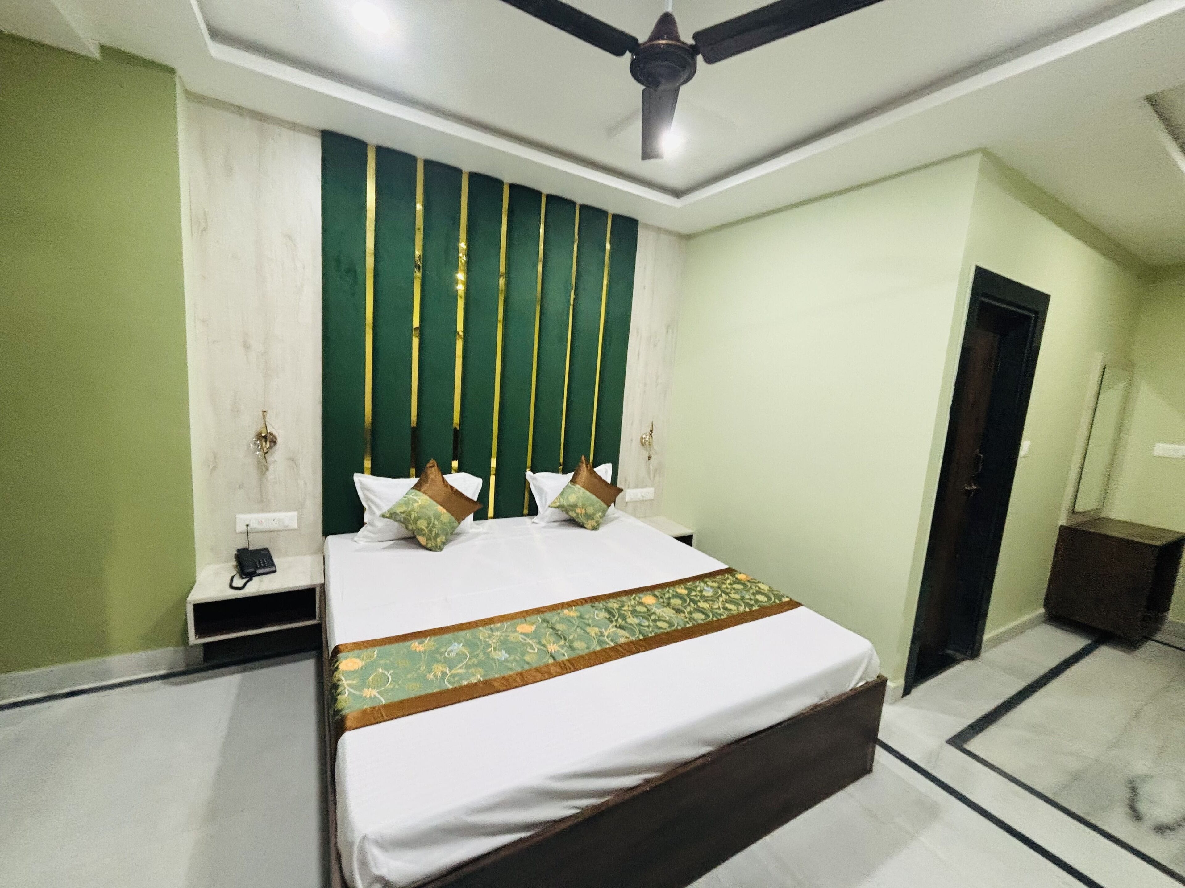Een hotelkamer met een bed, nachtkastje, telefoon, plafondventilator en een deur.