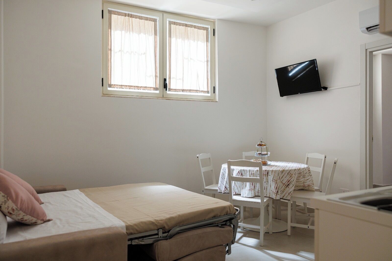 Una stanza piccola e pulita con un letto, un tavolo e sedie, una TV montata a parete e una finestra con tende.