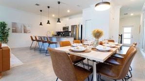 A modern kitchen with white cabinets, stainless steel appliances, and a dining area with a long table and brown chairs. - Stylish & Fun Retreat, Pool, Themed Rooms (Kissimmee)