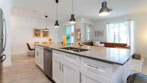 A modern kitchen with white cabinetry, a central island, pendant lighting, and a view to a living area with a sofa and curtains. - Stylish & Fun Retreat, Pool, Themed Rooms (Kissimmee)