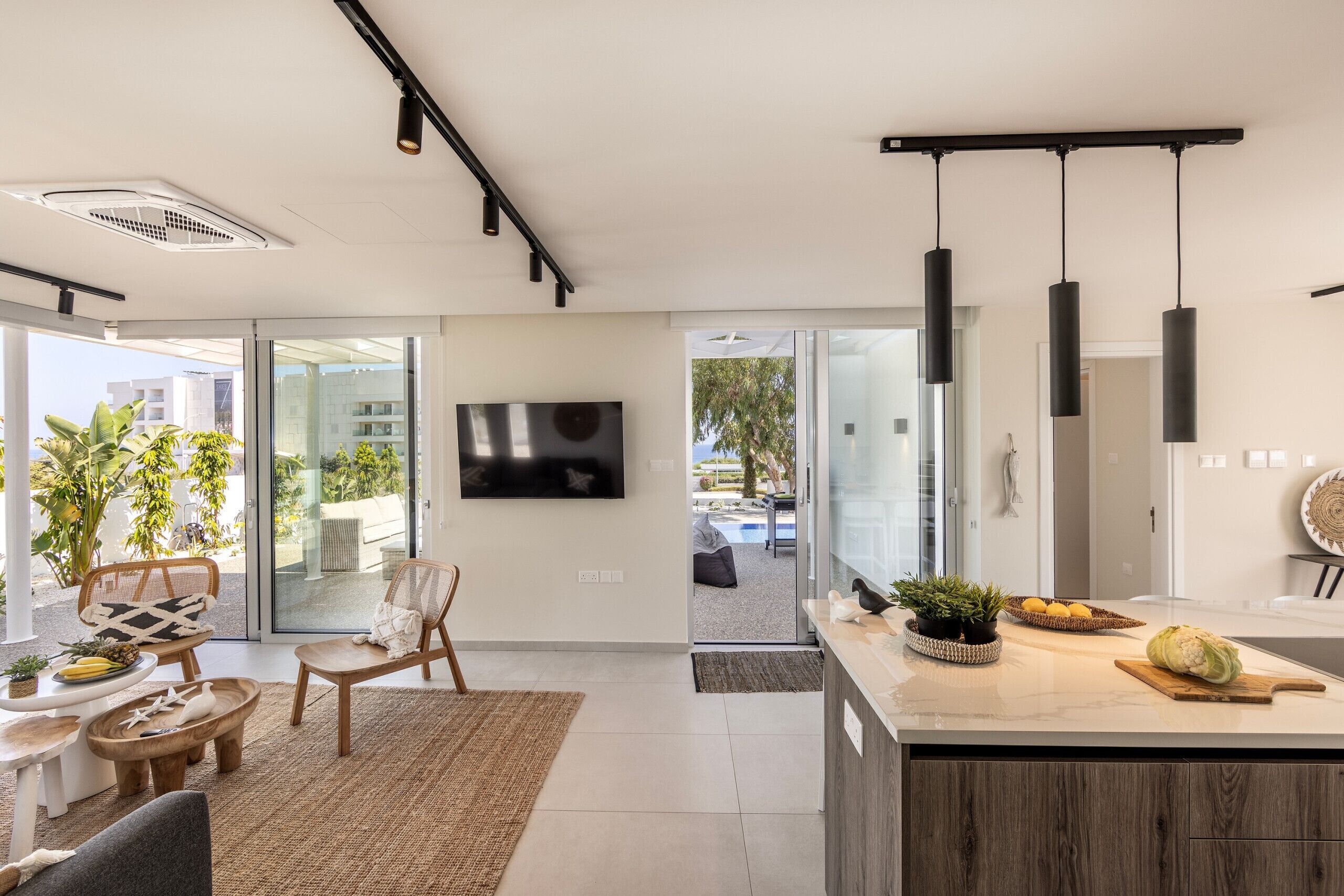 A modern living room with a large TV, comfortable seating, a kitchen area with a wooden island, and a view of the outdoors.