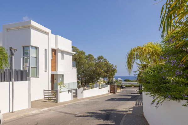 A modern white building with large windows and a wooden door, situated on a street with a clear view of the sea in the background.