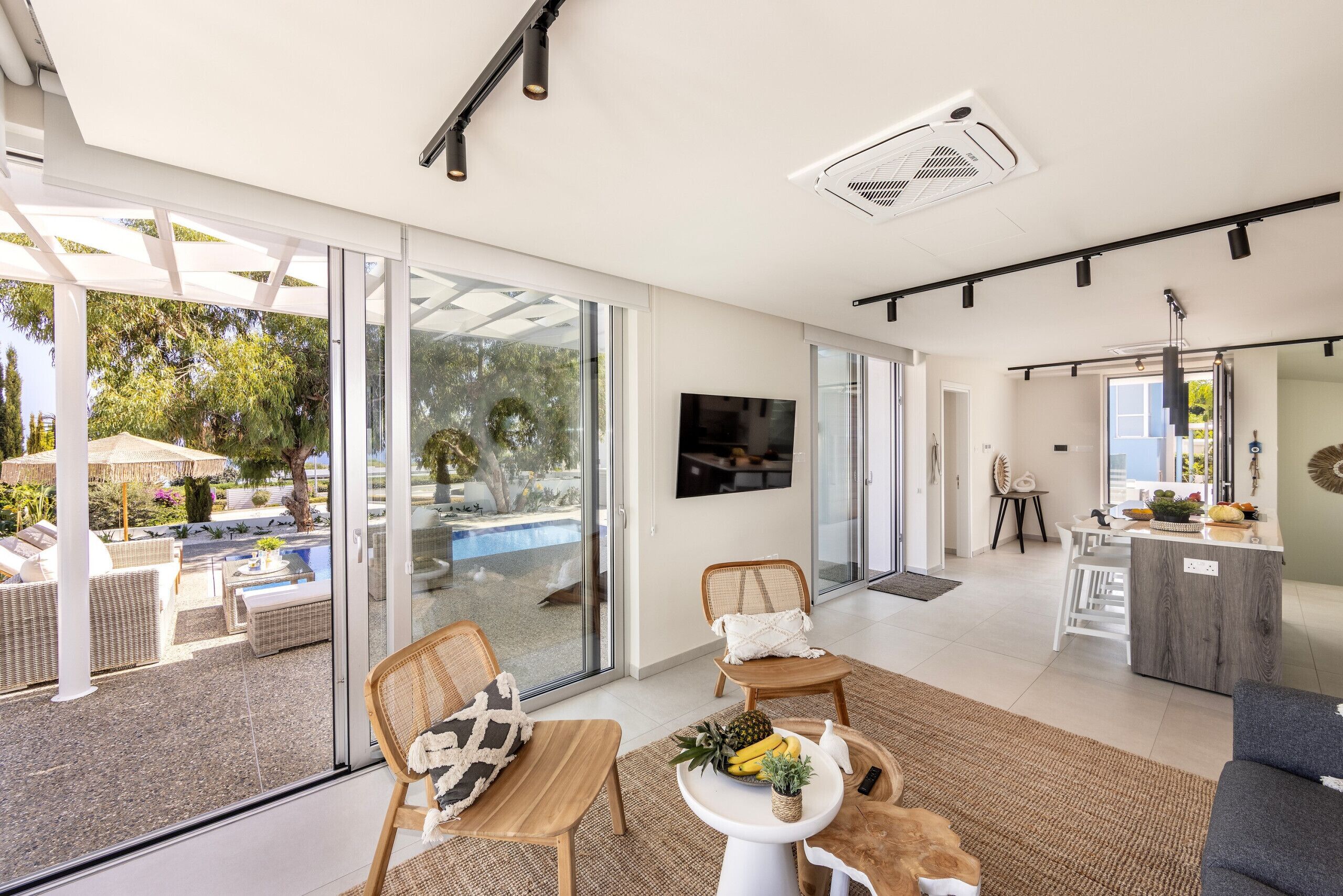 A spacious living area with a sliding glass door leading to a pool, wicker furniture, a small table with fruits, and a kitchen area with a bar.