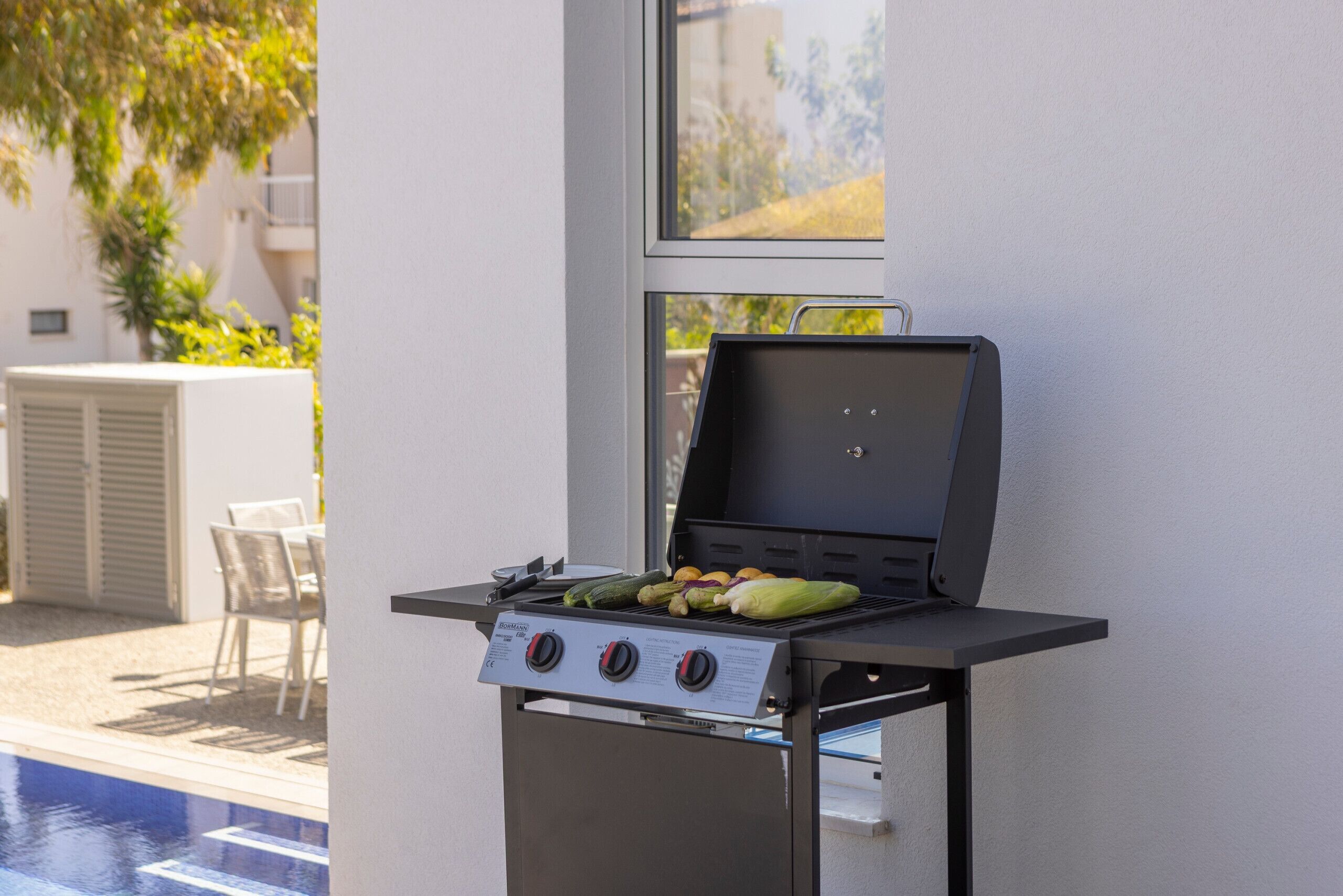 Een barbecue met groenten, geplaatst bij een raam met uitzicht op een zwembad en terras.