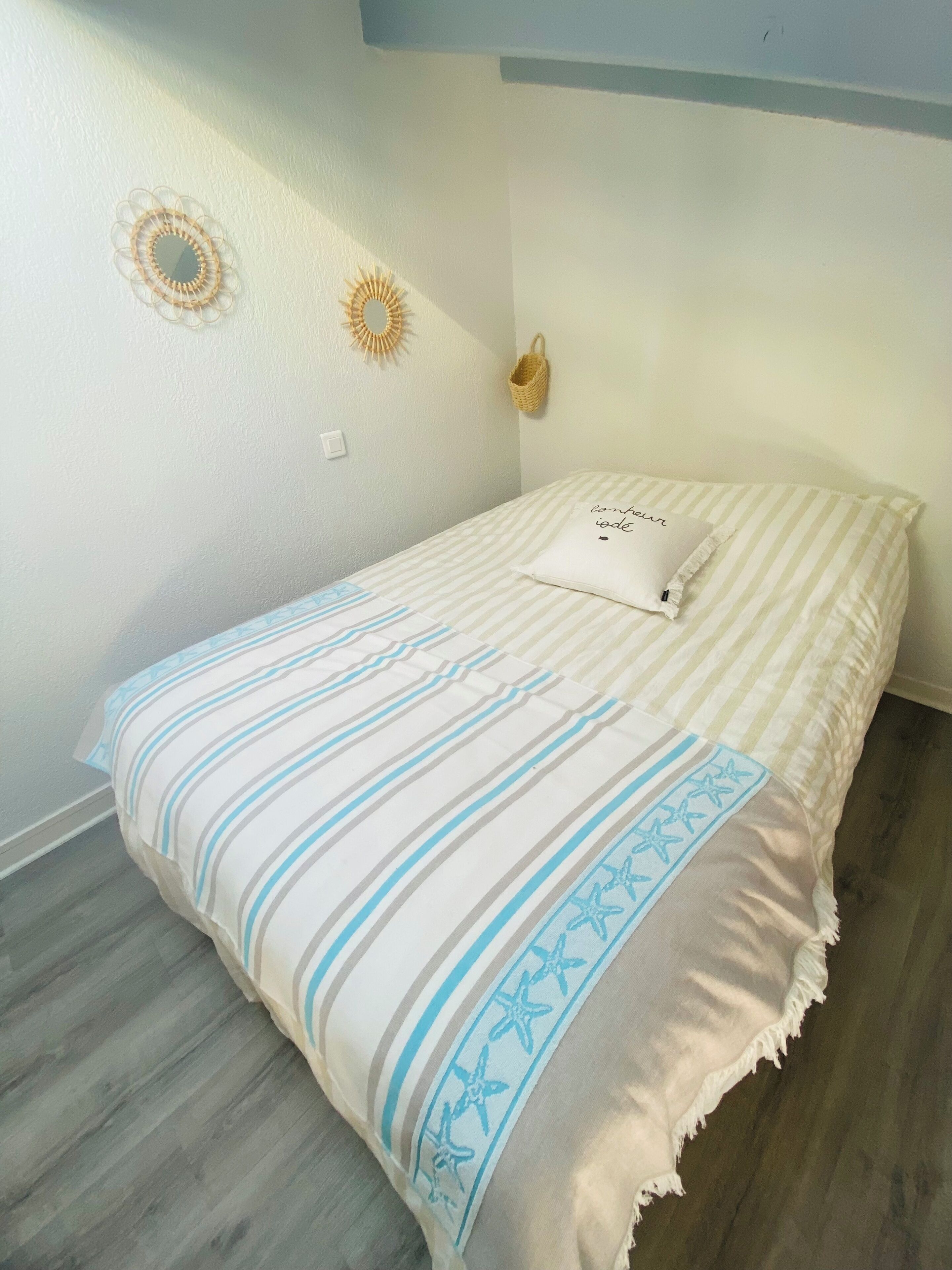 Ein ordentlich bezogenes Bett mit gestreifter Bettwäsche und einem Zierkissen, ein gewebter Wandbehang und ein kleiner Korb an der Wand.