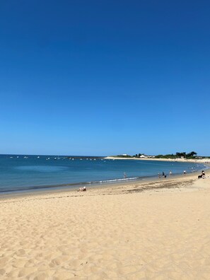 A wide sandy beach with clear blue water, a few boats in the distance, and a few people walking along the shore.