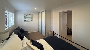 Een slaapkamer met een bed, een nachtkastje, een raam met jaloezieën, een schilderij en een deur naar een andere kamer.