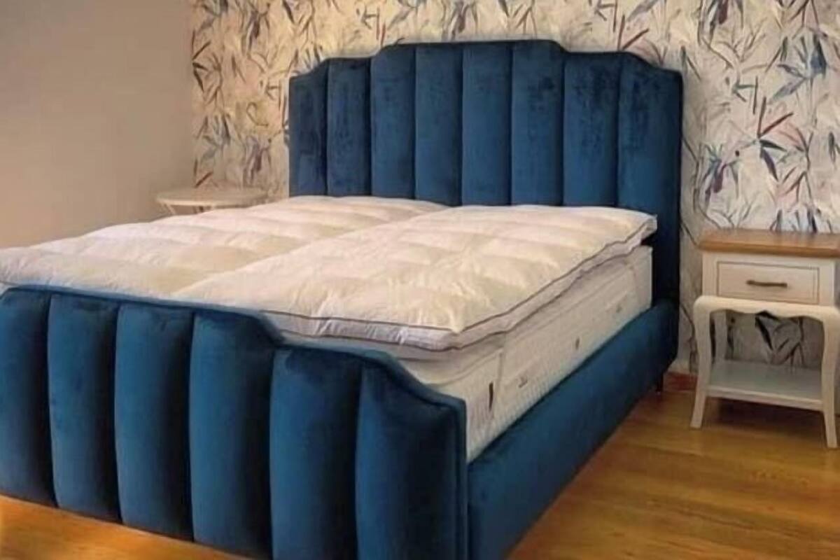 A bed with a blue upholstered headboard and white bedding in a room with patterned wallpaper and a wooden floor.