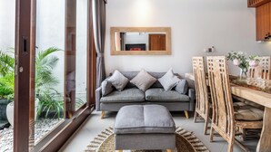A modern living room with a grey sofa, a wooden dining table, and a large mirror on the wall. - Private Pool Villa Dacira by Orivista (Canggu)