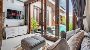 A modern living room with a flat-screen TV, a sofa, a coffee table, and a sliding glass door leading to an outdoor area with a pool. - Private Pool Villa Dacira by Orivista (Canggu)