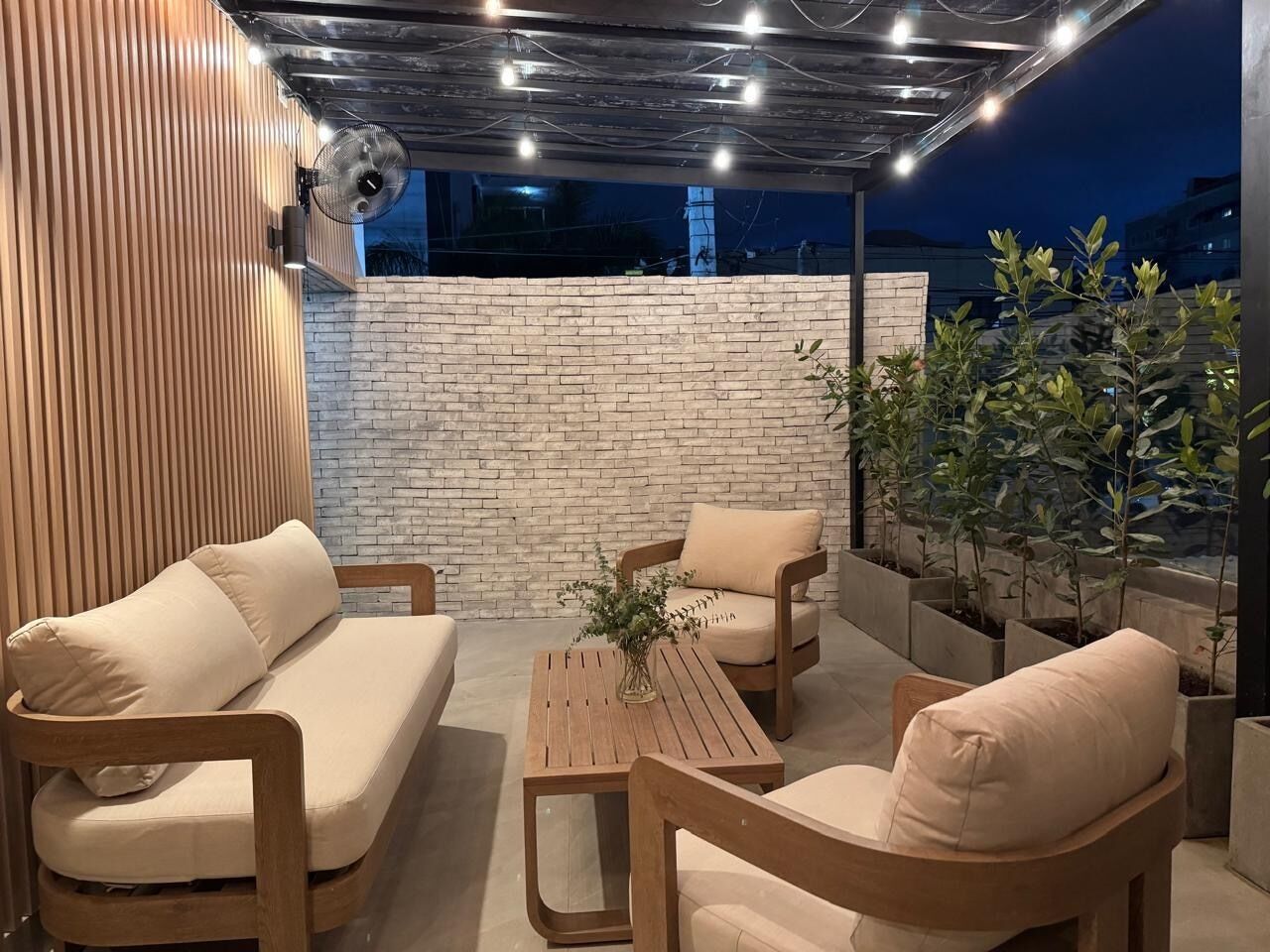 Un área de estar al aire libre con pared de ladrillo, mobiliario de madera y plantas en macetas.