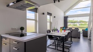 Une cuisine moderne dotée d’un îlot central, d’un coin repas avec une longue table et des chaises, et offrant une vue sur l’extérieur grâce à de grandes fenêtres.