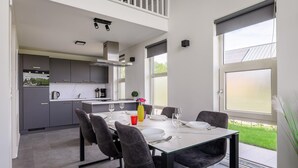 Une cuisine moderne avec des meubles gris, un coin repas doté d’une table en verre et une vue sur un jardin verdoyant grâce à de grandes fenêtres.