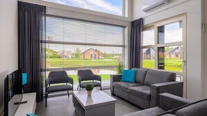 Un salon moderne comprenant un canapé, deux fauteuils, une table basse et un téléviseur. De grandes fenêtres offrent une vue sur un paysage verdoyant et des bâtiments.
