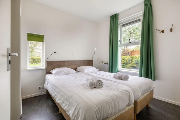 A bedroom with a bed, two windows with curtains, a bedside table, and a wall-mounted light.