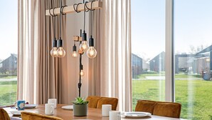 A dining area with a wooden table set for four, surrounded by chairs, with a view of a grassy yard and houses through large windows.