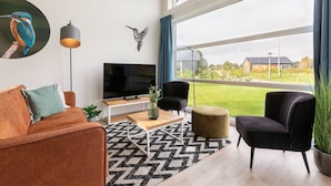 A modern living room with a sofa, a TV, a coffee table, and a large window with a view of a house and garden. - Tranquil villa for 6 guests with patio (Tynaarlo)