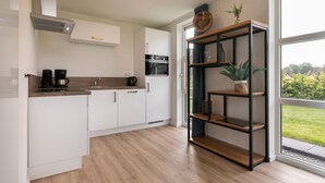 A modern kitchen with white cabinets, a built-in oven, and a coffee maker. There is a wooden shelf unit with plants and a window providing a view of greenery outside. - Tranquil villa for 6 guests with patio (Tynaarlo)