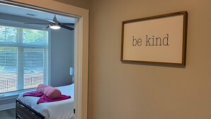 A bedroom with a bed, a ceiling fan, a window with blinds, and a wall with a framed sign saying 'be kind'.