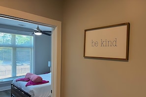 A bedroom with a bed, a ceiling fan, a window with blinds, and a wall with a framed sign saying 'be kind'.