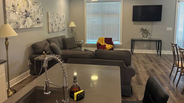 A modern living room with a sectional sofa, a dining area with a table and chairs, a kitchen with a bar area, and a flat-screen TV mounted on the wall.