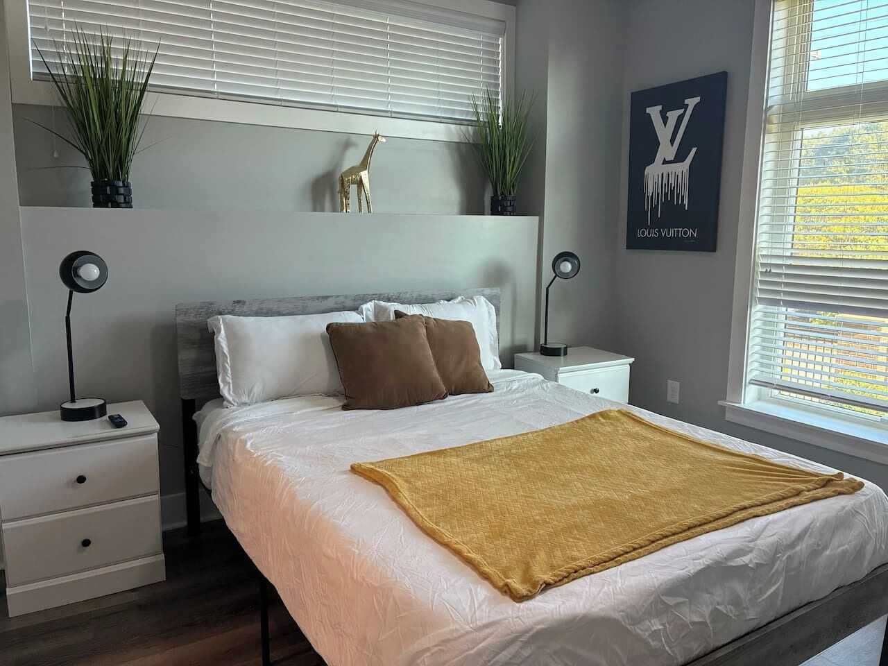 Un dormitorio moderno con una cama grande, ropa de cama blanca, un plaid color mostaza, un adorno de jirafa y un letrero de Louis Vuitton en la pared.