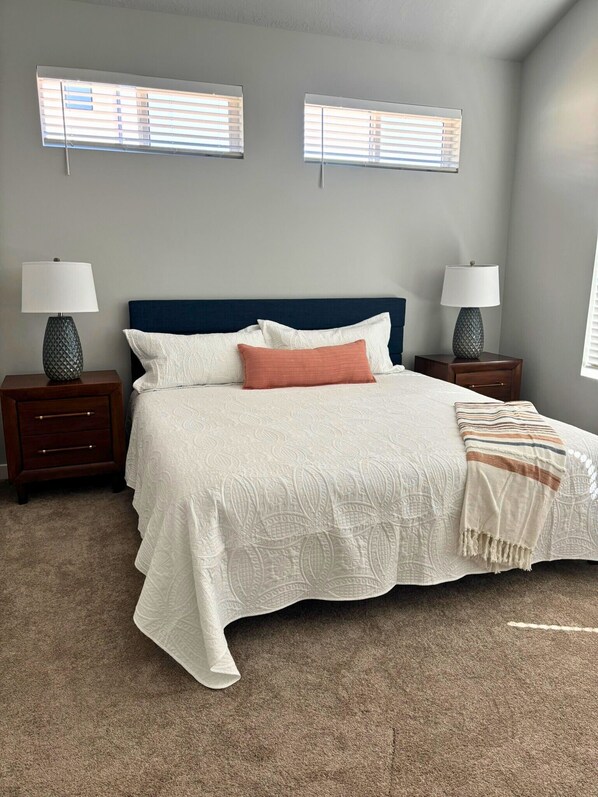 A bedroom with a bed, two nightstands, two lamps, and two windows with blinds.