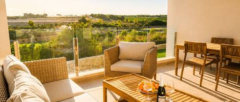A balcony with a wicker sofa, a wooden table with a bottle and glasses, and a dining set.