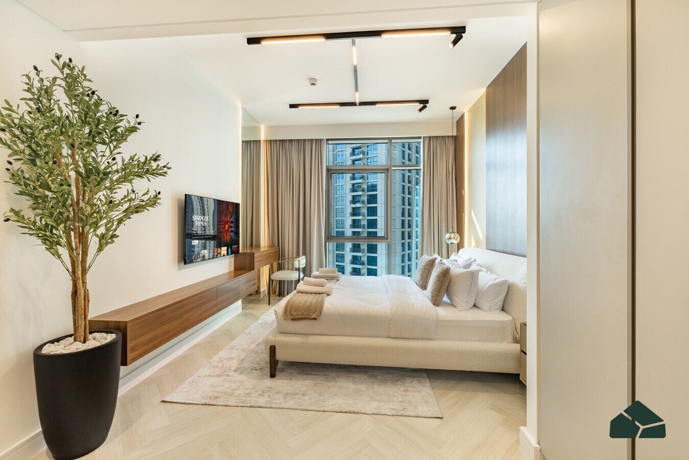 A modern hotel room with a large bed, a seating area, a potted plant, and a city view through the window.
