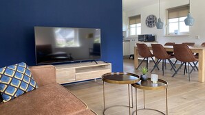 A modern living room with a large flat-screen TV, a brown sofa, and a dining area with a wooden table and chairs.