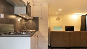 Eine kompakte Küche mit Gasherd, Spüle und Mikrowelle. Im angrenzenden Wohnbereich befindet sich eine Couch und ein Flachbildfernseher.