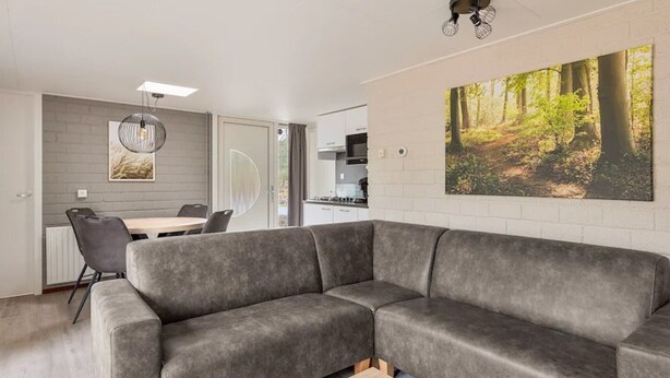 Ein modernes Wohnzimmer mit einem grauen Ecksofa, einem Esstisch mit Stühlen und einer Küche im Hintergrund.