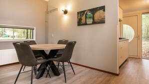 A dining area with a round table and four chairs, a painting on the wall, and a window with a view of trees. - Cozy Forest Bungalow in the Veluwe (Kootwijk)
