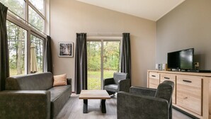 A modern living room with a sofa, armchairs, a coffee table, and a TV. Large windows offer a view of trees. - Cozy Forest Bungalow in the Veluwe (Kootwijk)