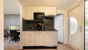 A modern kitchen with light wood cabinets, a black countertop, and a built-in microwave. There is a dining area with a table and chairs visible in the background. - Cozy Forest Bungalow in the Veluwe (Kootwijk)